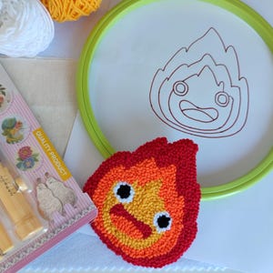 Könnte beinhalten: Ein Bastelprojekt mit einem fertigen rot-orange-gelben gestickten Flammendesign. Ein grüner Stickrahmen hält eine weiße Leinwand mit einer Flammenkontur. Bastelbedarf, Garn und Werkzeuge, mit einer Verpackung mit der Aufschrift "QUALITY PRODUCT".