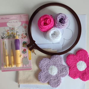 Op de afbeelding: Punch needle set met drie naalden, een hoepel, twee voorgemaakte bloemontwerpen en garen in roze, wit en paars.