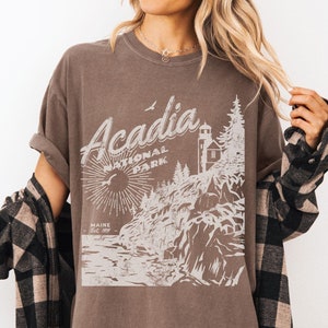 May include: A brown t-shirt with a vintage-style graphic of Acadia National Park, Maine. The graphic features a lighthouse, trees, and a sunburst.