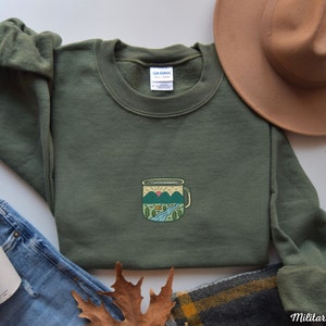 Może przedstawiać: Zielona bluza w kolorze khaki z grafiką przedstawiającą kubek do campingu z g&oacute;rskim krajobrazem w środku. Kubek jest obwiedziony czarną linią i wypełniony jasnobrązowym kolorem. G&oacute;ry są zielone i niebieskie z małym pomarańczowym słońcem wschodzącym nad nimi. W scenie znajduje się małe zielone drzewo i brązowy namiot.