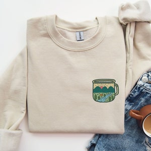 May include: A beige sweatshirt with a graphic of a camping mug with a mountain scene inside. The mug is green, blue, and orange. The sweatshirt is laid out on a white surface with a pair of blue jeans and a brown mug with coffee.