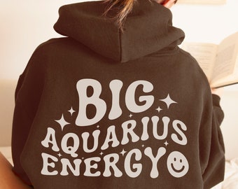 Puede incluir: Una sudadera con capucha marrón con el texto "BIG AQUARIUS ENERGY" en blanco, en una fuente de estilo retro. El texto está centrado en la parte posterior de la sudadera.