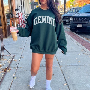 May include: A forest green sweatshirt featuring the word "GEMINI" in white. The crew neck sweatshirt is worn with white trainers and socks. The image is taken on a pavement. The colour of the sweatshirt is described as forest green.