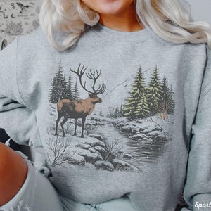 May include: A gray sweatshirt with a winter scene print featuring a reindeer standing in a snowy forest with a river in the background. The text "Sport Grey" is visible in the bottom right corner of the image.