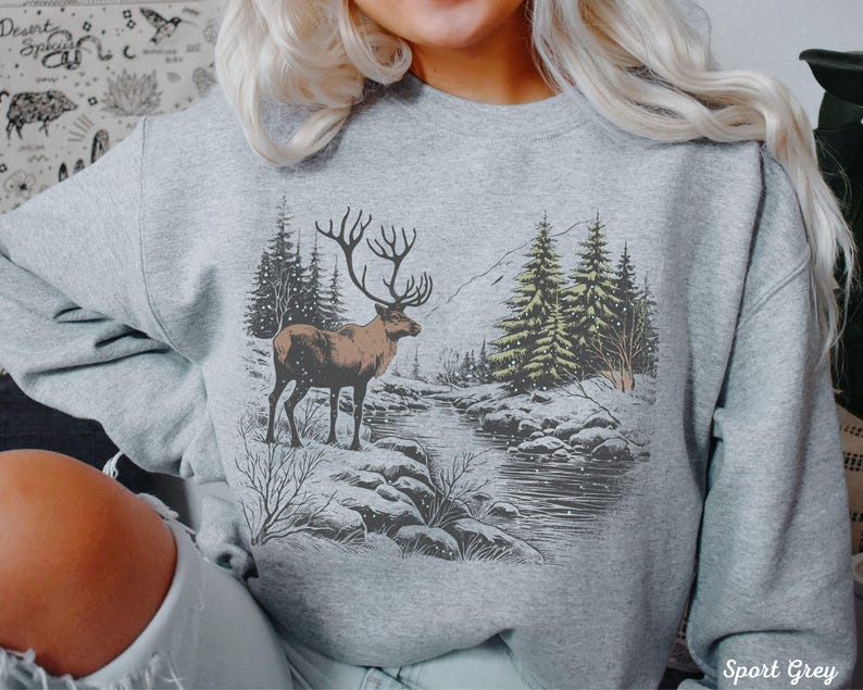 May include: A gray sweatshirt with a winter scene print featuring a reindeer standing in a snowy forest with a river in the background. The text "Sport Grey" is visible in the bottom right corner of the image.