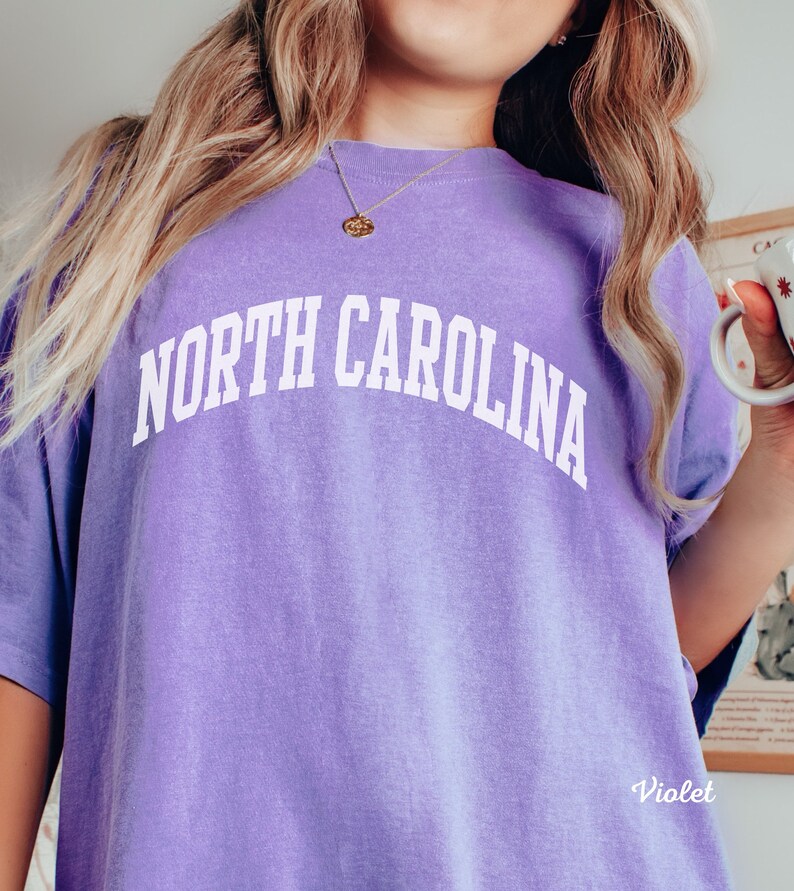 Może przedstawiać: Fioletowy t-shirt z napisem "NORTH CAROLINA" w kolorze białym, w łukowatych literach.