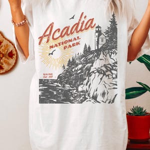 May include: White t-shirt with a vintage-style graphic of Acadia National Park, Maine. The graphic features a lighthouse, rocky cliffs, and a sun with rays. The text "Acadia National Park" is in red and the text "Maine Est. 1919" is in black.