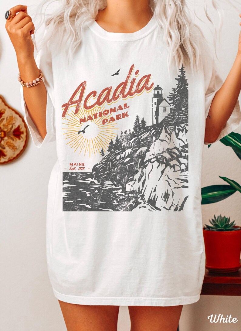 May include: White t-shirt with a vintage-style graphic of Acadia National Park, Maine. The graphic features a lighthouse, rocky cliffs, and a sun with rays. The text "Acadia National Park" is in red and the text "Maine Est. 1919" is in black.