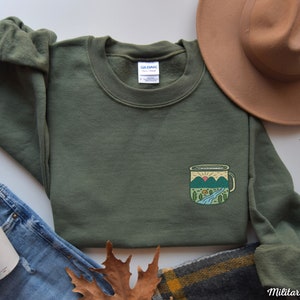 May include: A military green sweatshirt with a graphic of a camping mug with a mountain scene on the front. The mug has a sunrise, a river, and a small cabin in the scene. The sweatshirt is laid flat on a white surface with a brown hat, blue jeans, and a brown leaf.