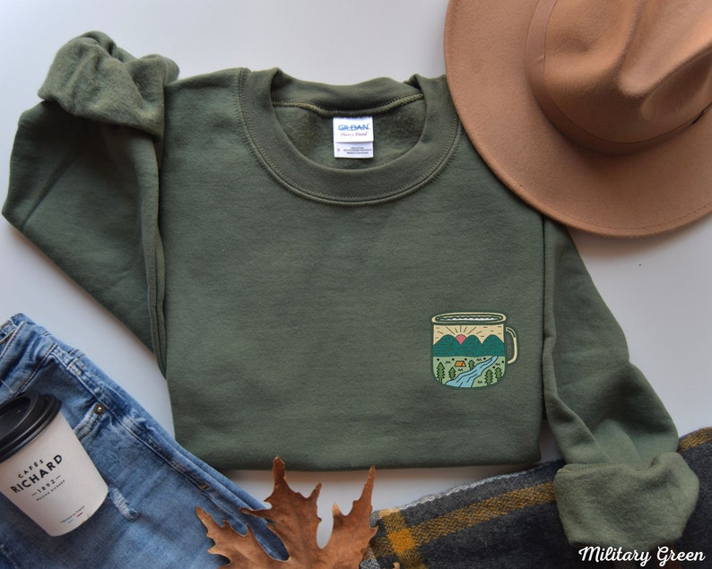 May include: A military green sweatshirt with a graphic of a camping mug with a mountain scene on the front. The mug has a sunrise, a river, and a small cabin in the scene. The sweatshirt is laid flat on a white surface with a brown hat, blue jeans, and a brown leaf.