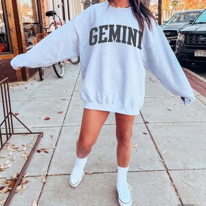 以下が含まれることがあります： グレーのトレーナーに、黒い文字で「GEMINI」とプリントされています。トレーナーはオーバーサイズでゆったりとしたフィット感です。モデルは白いスニーカーを履いています。