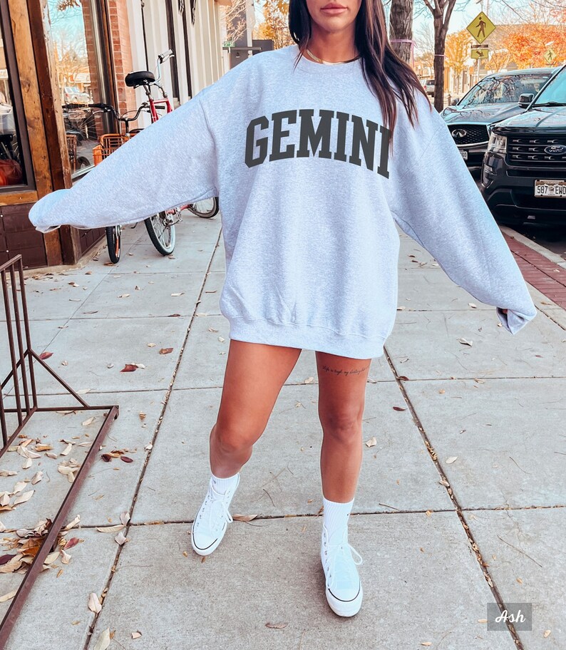 以下が含まれることがあります： グレーのトレーナーに、黒い文字で「GEMINI」とプリントされています。トレーナーはオーバーサイズでゆったりとしたフィット感です。モデルは白いスニーカーを履いています。