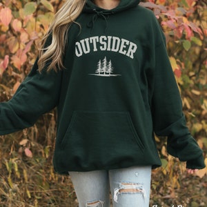 K&ouml;nnte beinhalten: Ein dunkelgr&uuml;ner Kapuzenpullover mit dem Wort "OUTSIDER" in wei&szlig;er Schrift &uuml;ber einer Grafik mit drei Kiefern. Der Pullover ist aus weichem, bequemem Material gefertigt und hat eine gro&szlig;e Tasche auf der Vorderseite.