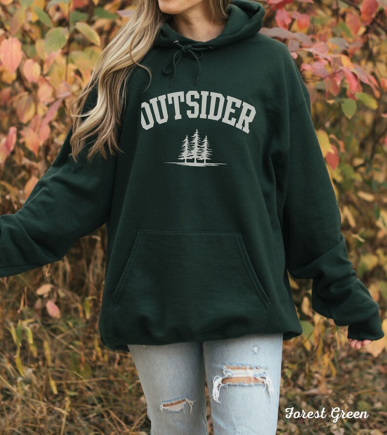 K&ouml;nnte beinhalten: Ein dunkelgr&uuml;ner Kapuzenpullover mit dem Wort "OUTSIDER" in wei&szlig;er Schrift &uuml;ber einer Grafik mit drei Kiefern. Der Pullover ist aus weichem, bequemem Material gefertigt und hat eine gro&szlig;e Tasche auf der Vorderseite.