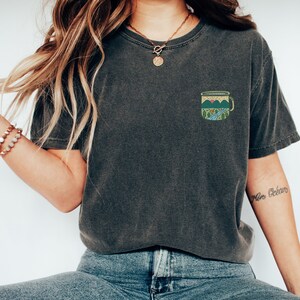 May include: A dark gray t-shirt with a small embroidered design of a camping mug with mountains and a sunset. The mug has a blue handle and a green interior. The t-shirt is worn by a person with long brown hair and a gold necklace.