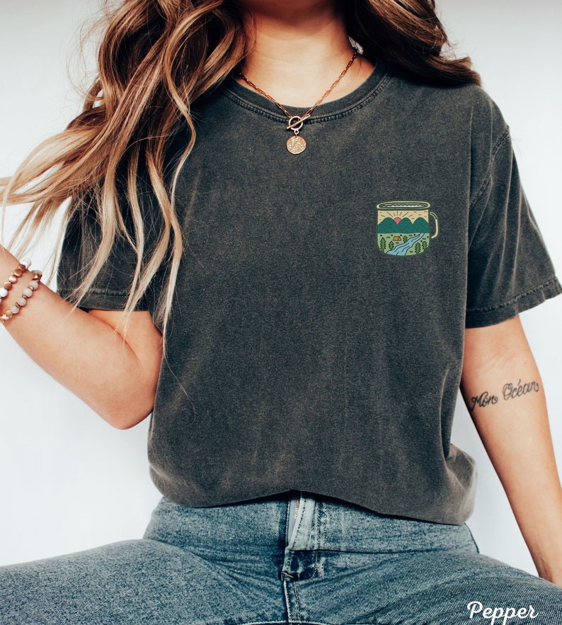 May include: A dark gray t-shirt with a small embroidered design of a camping mug with mountains and a sunset. The mug has a blue handle and a green interior. The t-shirt is worn by a person with long brown hair and a gold necklace.