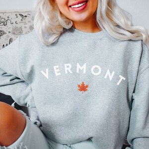 May include: A gray sweatshirt with the word "VERMONT" in white text and a red maple leaf. The sweatshirt is a crew neck style and is made of a soft, comfortable fabric. The color of the sweatshirt is Sport Grey.
