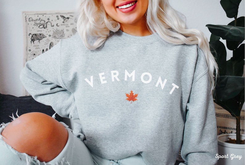 May include: A gray sweatshirt with the word "VERMONT" in white text and a red maple leaf. The sweatshirt is a crew neck style and is made of a soft, comfortable fabric. The color of the sweatshirt is Sport Grey.