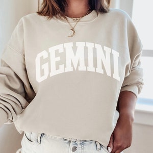 May include: A beige sweatshirt with the word "GEMINI" printed in large, white, arched letters. The sweatshirt has a crew neck and long sleeves. The person is wearing a gold necklace.