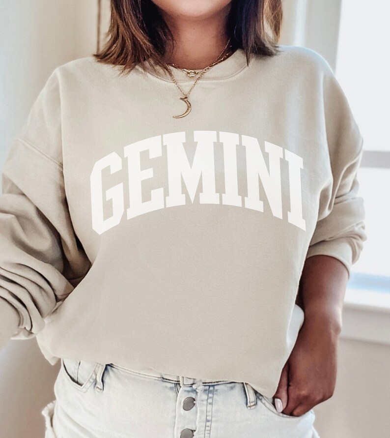 May include: A beige sweatshirt with the word "GEMINI" printed in large, white, arched letters. The sweatshirt has a crew neck and long sleeves. The person is wearing a gold necklace.