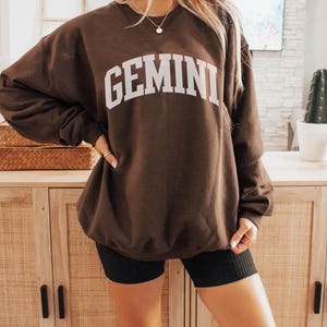 May include: A dark brown sweatshirt with the word "GEMINI" printed in white across the chest. The sweatshirt is a relaxed fit. The image has the word "Espresso" in the bottom right corner. The colour is similar to espresso coffee.