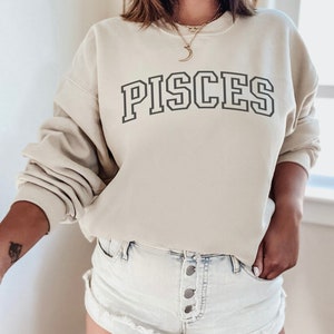 Pode incluir: Uma camisa de moletom bege claro com a palavra "PISCES" impressa em preto na frente. A camisa de moletom tem gola redonda e mangas compridas.
