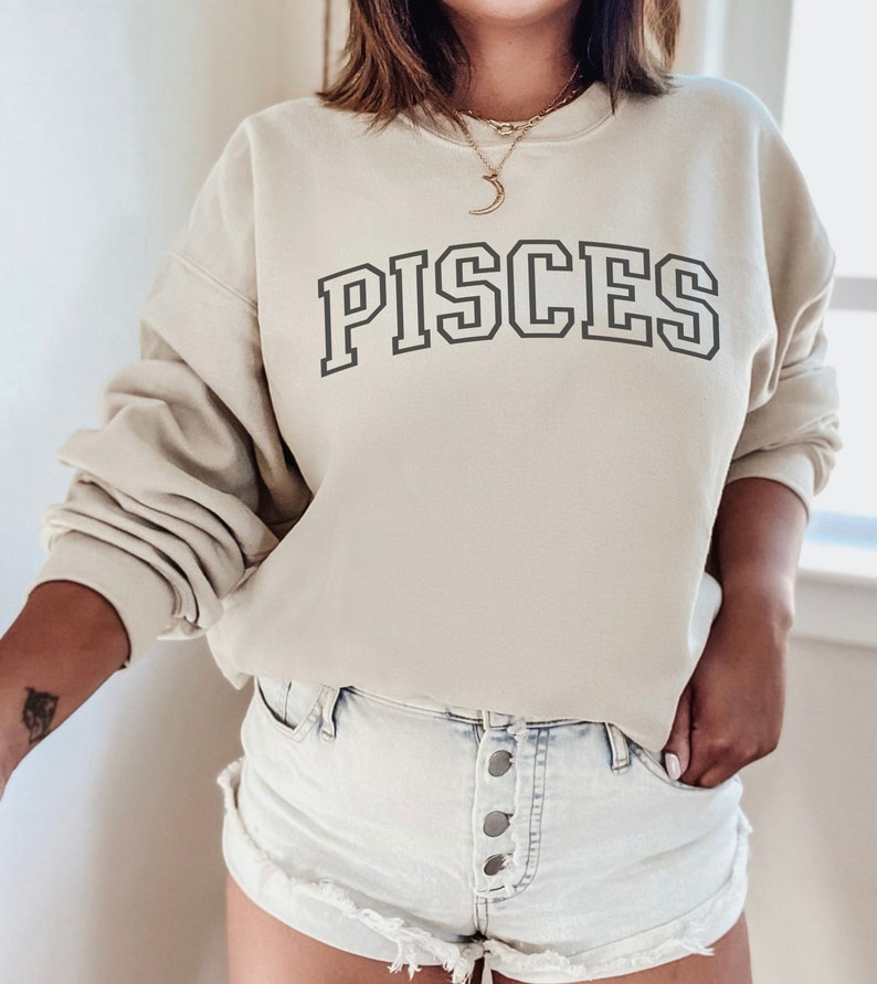 Pode incluir: Uma camisa de moletom bege claro com a palavra "PISCES" impressa em preto na frente. A camisa de moletom tem gola redonda e mangas compridas.