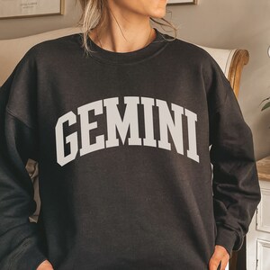 May include: Black sweatshirt featuring the word "GEMINI" in large, white, arched lettering. The crew neck sweatshirt is a dark colour. The text is in a bold, collegiate-style font.