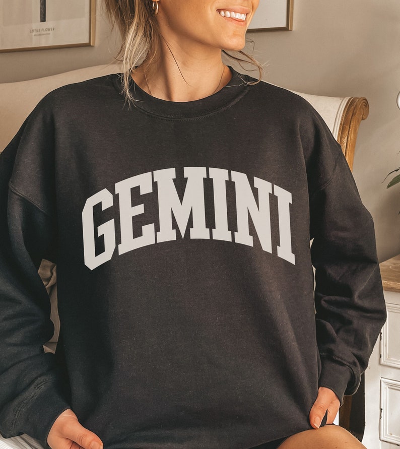 May include: Black sweatshirt featuring the word "GEMINI" in large, white, arched lettering. The crew neck sweatshirt is a dark colour. The text is in a bold, collegiate-style font.