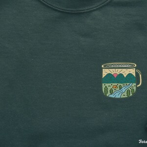 May include: Forest green sweatshirt with a graphic of a camping mug with a mountain scene and a sunrise. The mug has a white rim and handle.