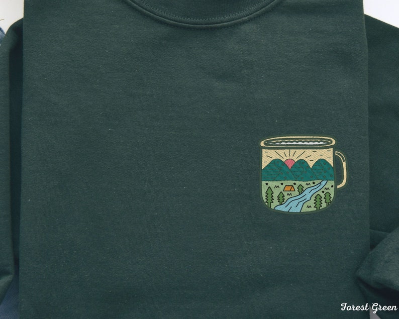 May include: Forest green sweatshirt with a graphic of a camping mug with a mountain scene and a sunrise. The mug has a white rim and handle.