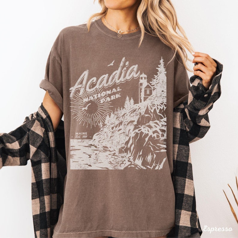 May include: A brown t-shirt with a vintage-style graphic of Acadia National Park, Maine. The graphic features a lighthouse, trees, and a sunburst.