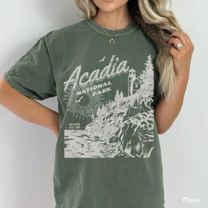 May include: A green t-shirt with a white graphic design of Acadia National Park, Maine. The design features a lighthouse, trees, and a rocky coastline. The text "Acadia National Park" and "Maine Est. 1919" are included in the design.