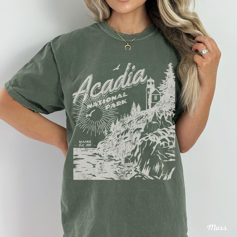 May include: A green t-shirt with a white graphic design of Acadia National Park, Maine. The design features a lighthouse, trees, and a rocky coastline. The text "Acadia National Park" and "Maine Est. 1919" are included in the design.