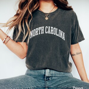 Może przedstawiać: Szary t-shirt z napisem "NORTH CAROLINA" w białych literach. Koszulkę nosi osoba z długimi brązowymi włosami i złotym naszyjnikiem z okrągłym wisiorkiem.