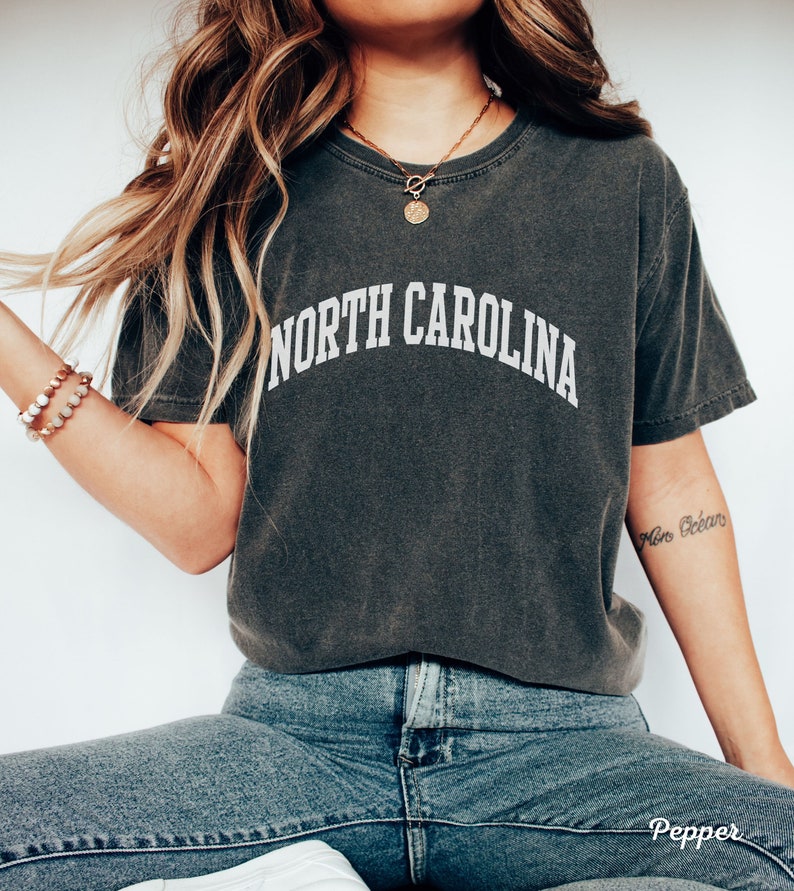 Może przedstawiać: Szary t-shirt z napisem "NORTH CAROLINA" w białych literach. Koszulkę nosi osoba z długimi brązowymi włosami i złotym naszyjnikiem z okrągłym wisiorkiem.