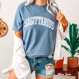 May include: A blue t-shirt with the word "SAGITTARIUS" printed in white on the front. The shirt is worn by a person wearing blue denim shorts.
