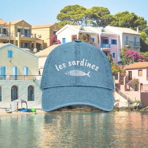 May include: A blue denim baseball cap with white embroidery that reads "les sardines" and a white embroidered fish.