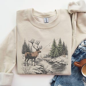 May include: A beige sweatshirt with a graphic print of a reindeer standing in a snowy forest scene. The reindeer is brown with large antlers. The forest is green and brown with a small stream running through it.