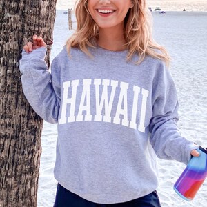 May include: A gray sweatshirt with the word "HAWAII" printed in white on the front. The sweatshirt is being worn by a person standing on a beach.