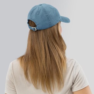 May include: A blue denim baseball cap with a buckle closure. The cap is worn backwards on a person with long brown hair.