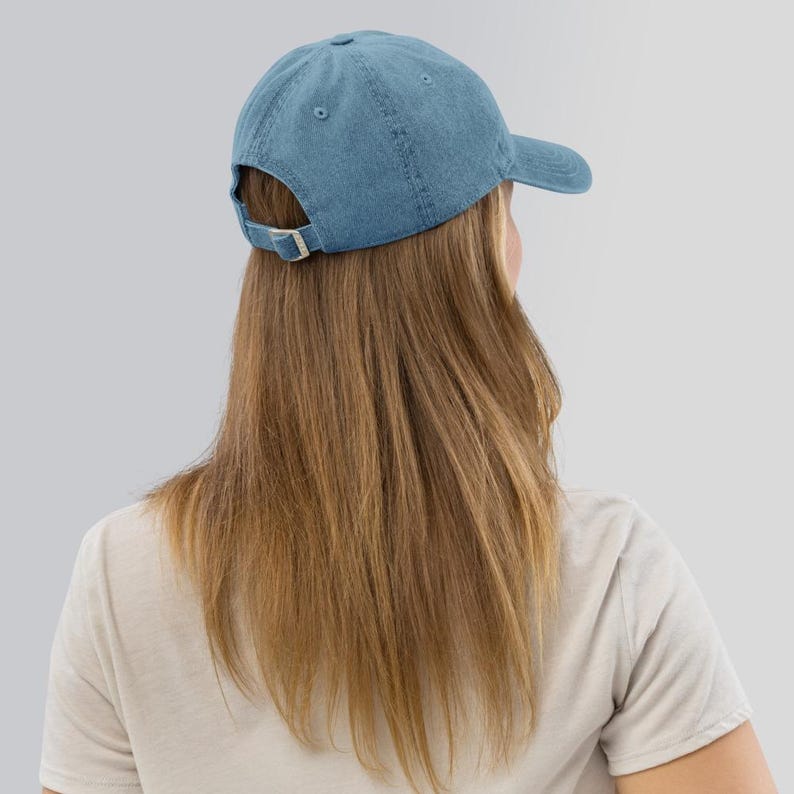 May include: A blue denim baseball cap with a buckle closure. The cap is worn backwards on a person with long brown hair.
