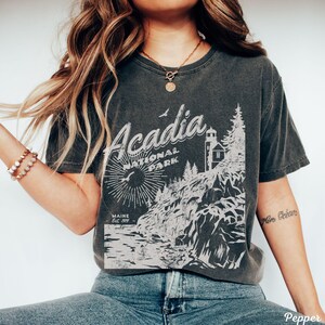 May include: A dark gray t-shirt with a vintage-style graphic of Acadia National Park in Maine. The graphic features a lighthouse, trees, and a rocky coastline. The text "Acadia National Park" and "Maine Est. 1919" is printed on the shirt.