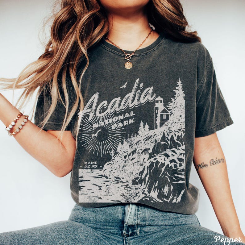 May include: A dark gray t-shirt with a vintage-style graphic of Acadia National Park in Maine. The graphic features a lighthouse, trees, and a rocky coastline. The text "Acadia National Park" and "Maine Est. 1919" is printed on the shirt.