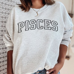 Pode incluir: Camisola de moletom cinza com a palavra "PISCES" impressa em letras pretas com contorno.
