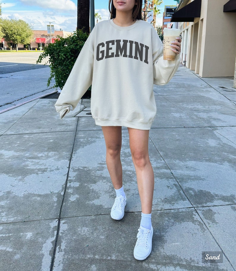 以下が含まれることがあります： クリーム色のスウェットシャツに、前面に茶色の「GEMINI」という文字がプリントされています。スウェットシャツは、歩道に立っている人が着用しています。