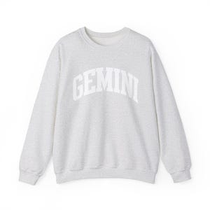 May include: A light grey sweatshirt featuring the word "GEMINI" in white, arched lettering. The crew neck sweatshirt has long sleeves and a speckled fabric texture. The design is simple and modern.