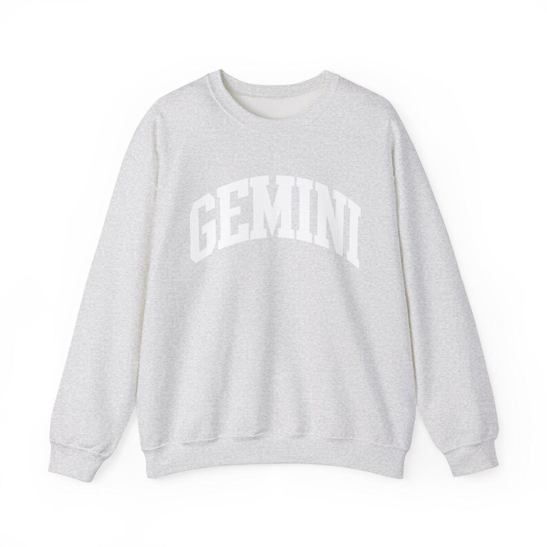 May include: A light grey sweatshirt featuring the word "GEMINI" in white, arched lettering. The crew neck sweatshirt has long sleeves and a speckled fabric texture. The design is simple and modern.