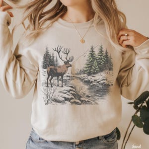 May include: A beige sweatshirt with a graphic print of a reindeer standing in a snowy forest scene. The reindeer is brown with large antlers. The forest is green and white with a river running through it.