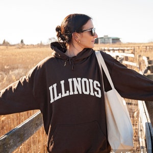 May include: A brown hoodie with the word "ILLINOIS" in white lettering. The hoodie is worn by a person standing on a wooden walkway. The background is a field of tall grass.